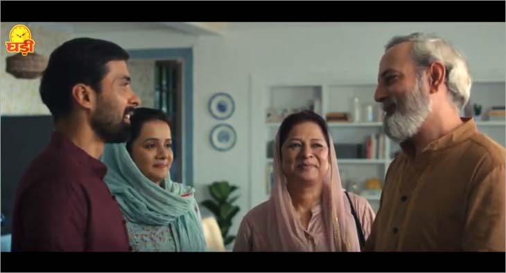 A young man greets an older couple at home while a woman stands beside him, all smiling warmly in a welcoming family setting.