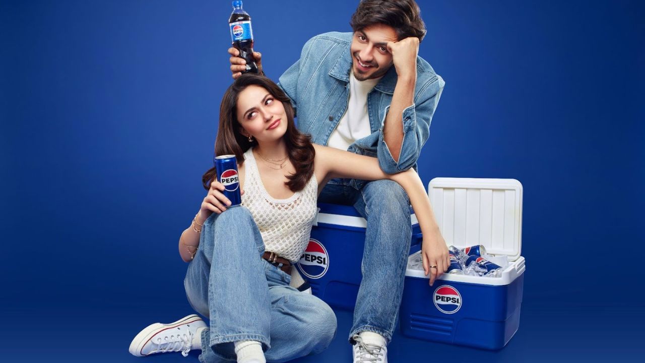 A young couple sits casually beside a cooler filled with Pepsi bottles, playfully highlighting the drink, with the man holding a bottle above the woman’s head to suggest “extra fizz.”