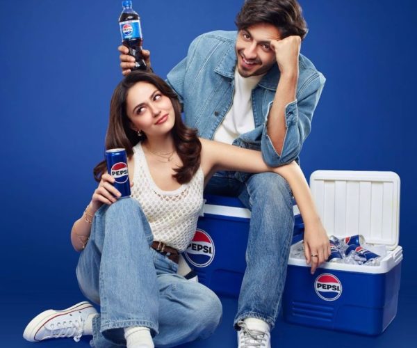 A young couple sits casually beside a cooler filled with Pepsi bottles, playfully highlighting the drink, with the man holding a bottle above the woman’s head to suggest “extra fizz.”