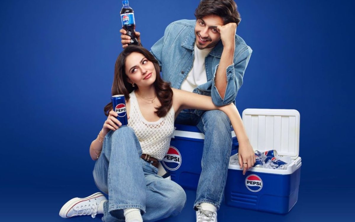 A young couple sits casually beside a cooler filled with Pepsi bottles, playfully highlighting the drink, with the man holding a bottle above the woman’s head to suggest “extra fizz.”