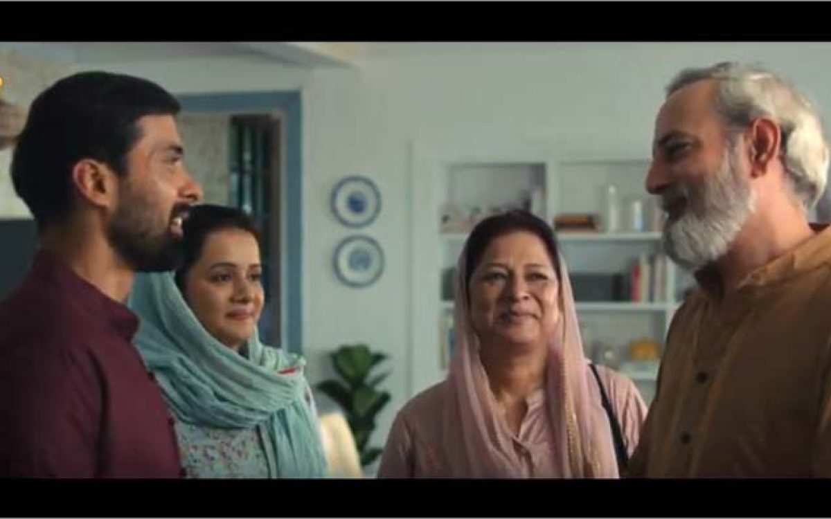 A young man greets an older couple at home while a woman stands beside him, all smiling warmly in a welcoming family setting.
