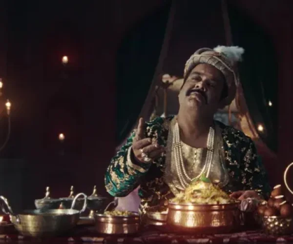A royal-looking man in traditional attire sits at a lavish dining table enjoying biryani, highlighting India Gate’s humorous take on the “royal biryani” narrative.